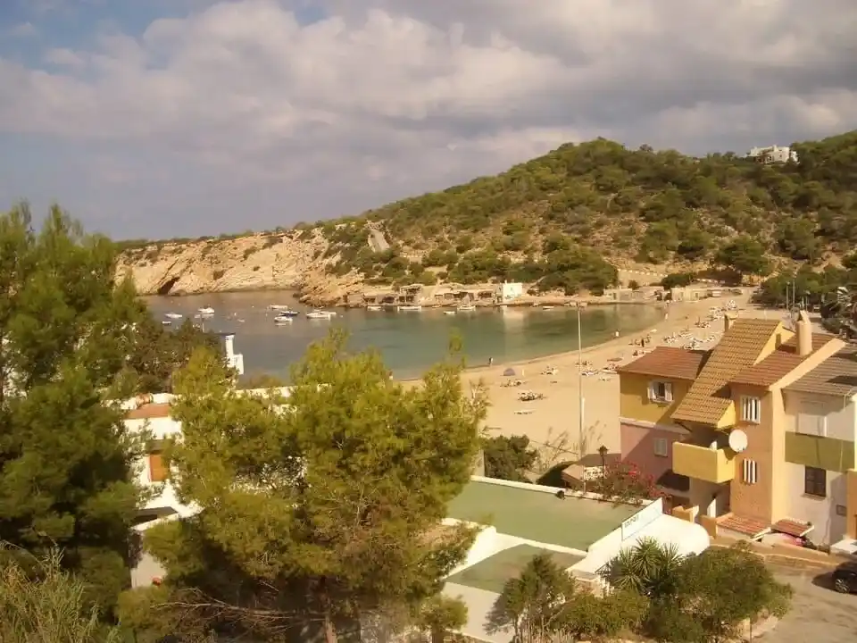 Vista panorámica de la playa de Cala Vadella con aguas turquesas, barcos fondeados y colinas boscosas de fondo.