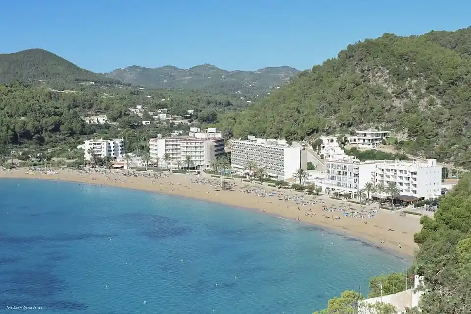 Vista panorámica de la playa de Cala San Vicente en Ibiza con aguas cristalinas, hoteles a pie de playa y colinas de pinos