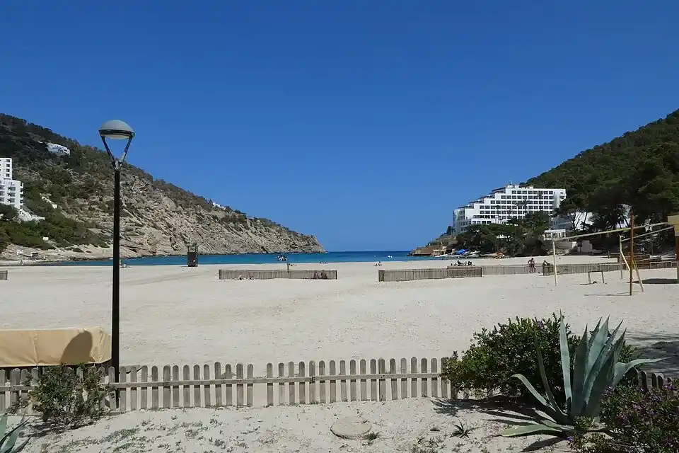 Panorámica de la amplia playa de arena de Cala Llonga entre colinas verdes y complejos hoteleros en Ibiza