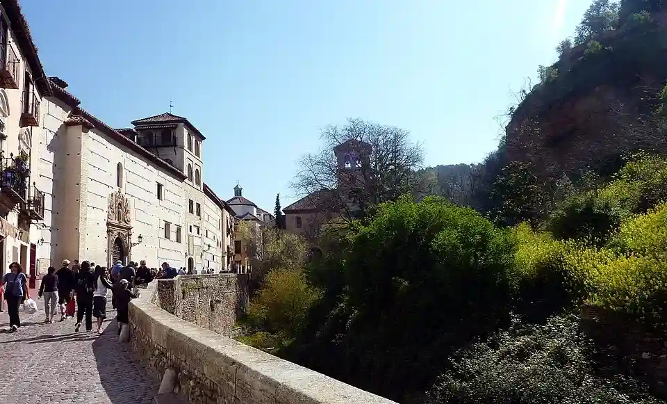 Peatones recorriendo la empedrada Carrera del Darro junto al Convento de Zafra y la ribera del río hacia el Paseo de los Tristes en Granada.