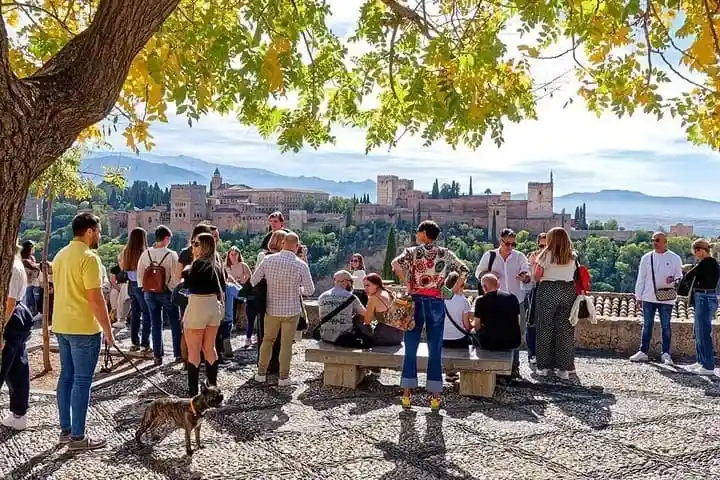 Turistas en la terraza empedrada del Mirador de San Nicolás contemplando las vistas panorámicas de la Alhambra y Sierra Nevada en Granada.