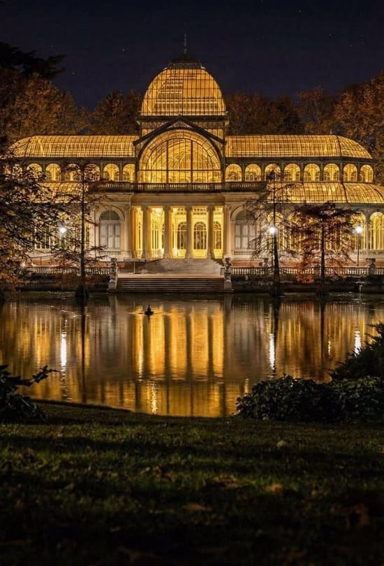 Fachada del Palacio de Cristal del Retiro iluminada de noche con su reflejo en el lago de Madrid