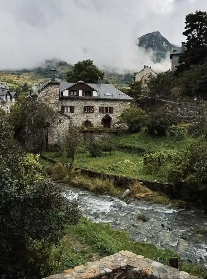 Casas de piedra tradicionales y montañas nubladas junto al río, un paisaje imprescindible que ver en Sallent de Gállego.