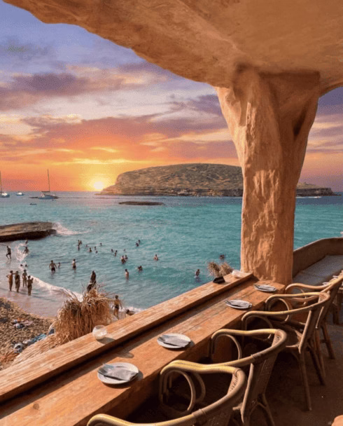 Vista panorámica del atardecer en Cala Comte, Ibiza, desde la terraza de un restaurante con arcos de piedra. Se observa el mar turquesa, la isla s'Illa des Bosc al fondo y el cielo con colores naranjas y violetas.