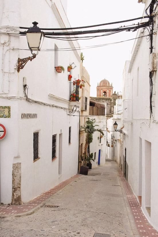 strecha calle encalada del casco antiguo de Tarifa con casas blancas tradicionales, farolas antiguas y la torre de una iglesia visible al fondo entre los cables eléctricos