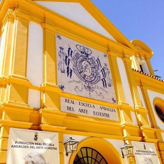 Fachada amarilla de la Real Escuela Andaluza del Arte Ecuestre en Jerez con escudo real y cartel institucional
