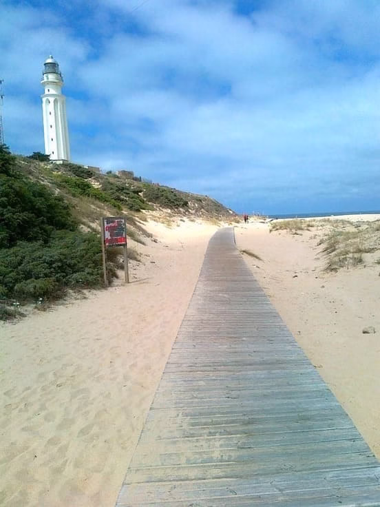 Pasarela de madera sobre dunas de arena que conduce al Faro de Trafalgar con el cielo azul y el mar Atlántico al fondo en la playa de Caños de Meca, Cádiz