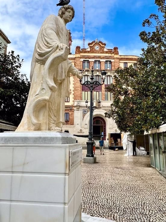 Estatua de mármol en la Plaza de San Antonio de Cádiz con el histórico Palacio de la Aduana al fondo, pavimento de adoquines tradicional