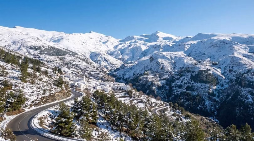 Carretera de montaña que llega a un pueblo blanco rodeado de montañas nevadas en Sierra Nevada, uno de los mejores sitios de montaña en Andalucía para disfrutar de la nieve